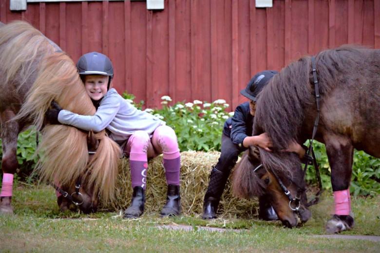 Kaksi lasta istuu heinäpaalilla, kummatkin halaavat ponejaan.