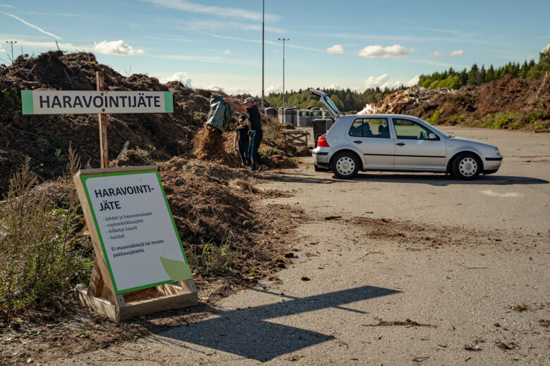 Henkilöauto pysäköitynä jäteaseman haravointijätteen kokoamispaikalle. Mies tyhjentää säkistä haravaointijätettä kasaan. Lapsi seuraa vieressä.
