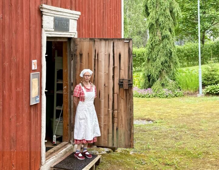 Tarvasjoen kotiseutumuseon punainen puurakennus ja ovella perinteiseen asuun pukeutunut opas.