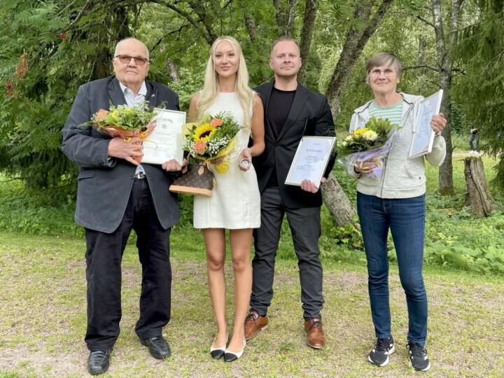 Neljä henkiöä seisoo rinnakkain vehreässä maisemassa ja pitävät kädessään kukkia, Lieto-mitalia ja kunniakirjoja.