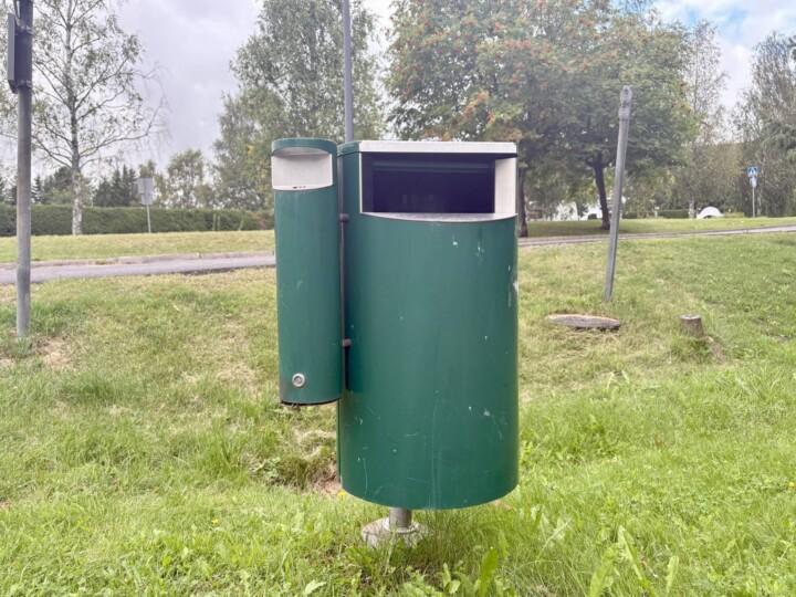 Vihreä roskapönttö, jonka kyljessä erillinen tumppiroskis. Taustalla oja ja tie.